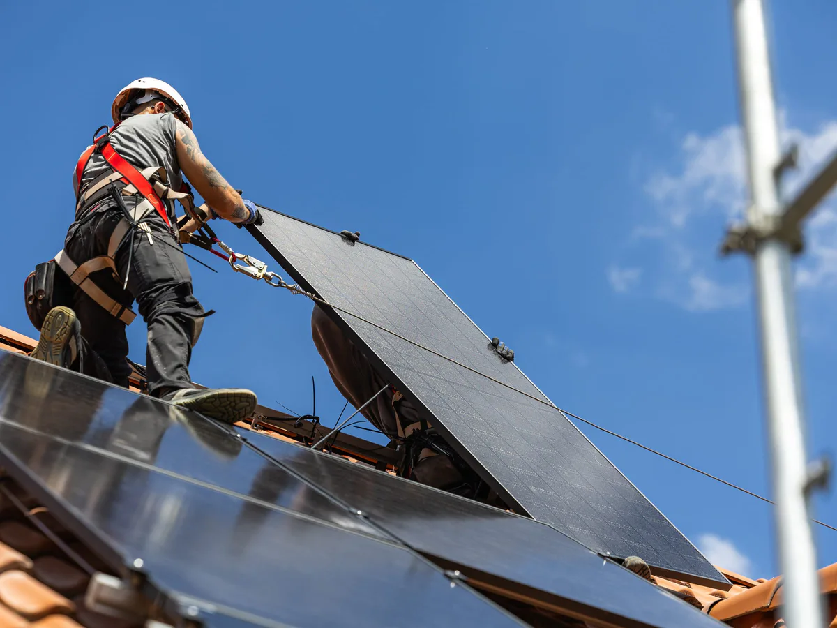 Monteur mit Sicherheitsgurt montiert ein Solarmodul auf einem Ziegeldach